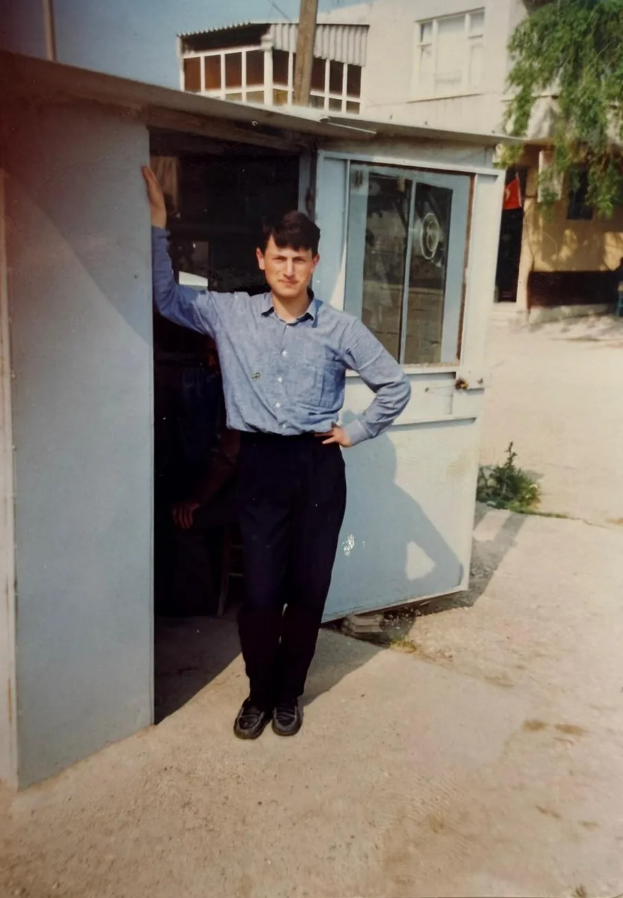 Genç bir adam, açık mavi dükkanın kapısının önünde elini kapıya dayamış duruyor.