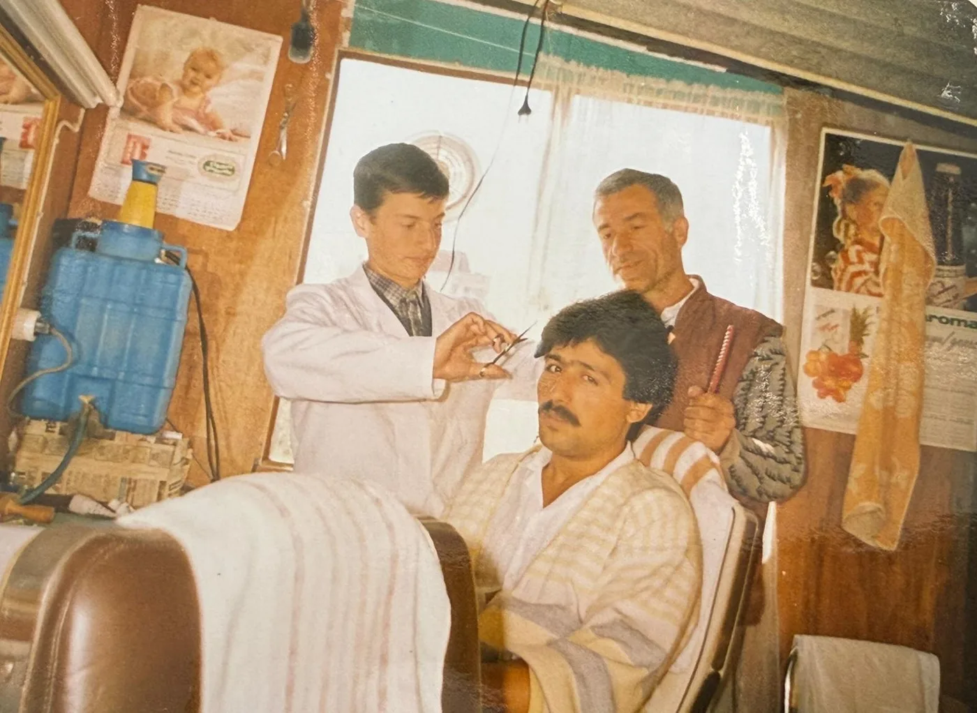 Dükkanın içinde genç bir berber makasla saç kesiyor, arkasında elinde tarak tutan bir berber işi izliyor.